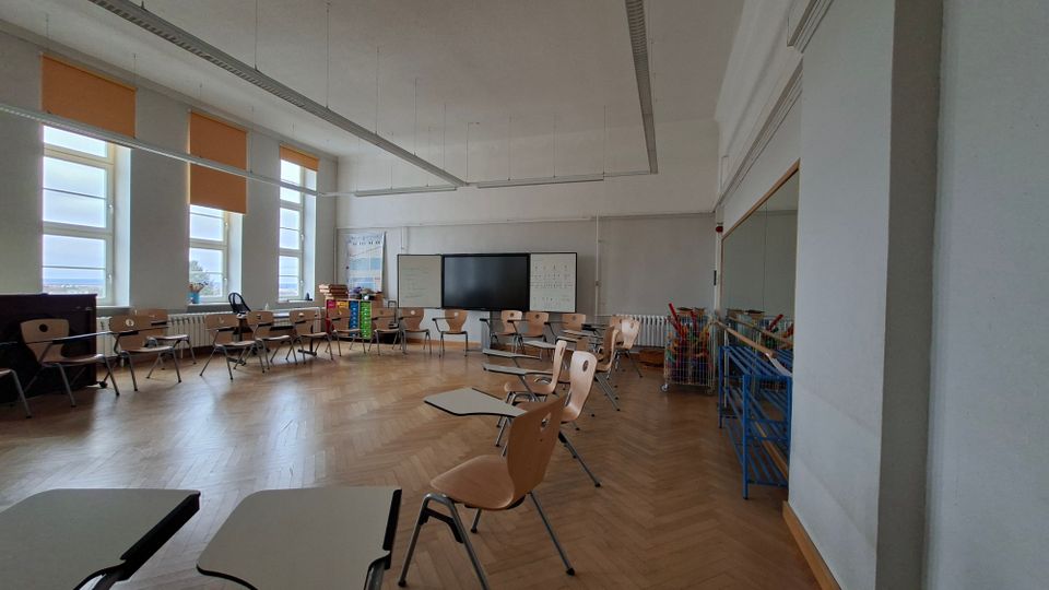 Heller Klassenraum mit großen Fenstern und orangefarbenen Rollos, Parkettboden und in einem Halbkreis angeordneten Stühlen mit Schreibflächen. Vorn stehen ein großer Bildschirm zwischen Whiteboards sowie Regale und Materialwagen an der Wand.