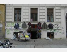 Fassade eines Altbaus mit einem Eingang zu einem Geschäft im Untergeschoss, vor dem Kleidung aufgestellte Schaufensterpuppen und Kleidungsstücke präsentiert werden. Die Wand ist mit Graffiti besprüht, und davor stehen einige Stühle und Kleinteile auf dem Gehweg.