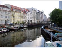 Reihe historischer Gebäude mit pastellfarbenen Fassaden entlang eines Kanals, an dem mehrere kleinere Schiffe und Boote festgemacht sind; das Ufer ist mit Gehwegen und Geländern gestaltet, umrahmt von Bäumen auf der rechten Seite.