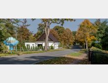 Straße mit Herbstbäumen in einer ländlichen Umgebung, flankiert von einem traditionellen Fachwerkhaus mit einem großen Baum davor. Links steht ein Willkommensschild mit der Aufschrift „Lindow (Mark)“, das die Ortschaft markiert.