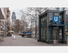 Straßenszene in Berlin mit einem gläsernen Eingang zur U-Bahn-Station Uhlandstraße rechts im Bild; im Hintergrund ist die Kaiser-Wilhelm-Gedächtniskirche zu erkennen. Links befinden sich Geschäfte, Restaurants und eine breite Gehwegpassage.