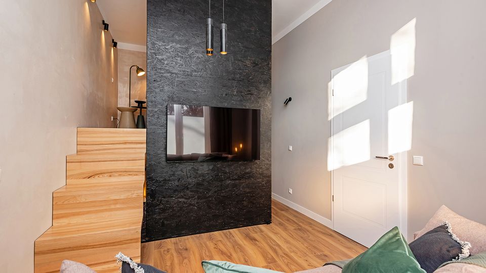 Modernes Wohnzimmer mit Holzfußboden und minimalistischer Einrichtung, dominiert von einer schwarzen Akzentwand mit integriertem Flachbildfernseher und hängenden Pendelleuchten. Links führt eine helle Holztreppe zu einer erhöhten Nische, rechts befindet sich eine weiße Tür in einer grauen Wand mit einfallendem Sonnenlicht.