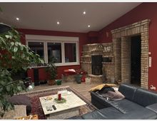 Wohnzimmer mit roten Wänden, großer Fensterfront und hellbraunem Holzfußboden. Ein gemauerter Kamin mit Regalen aus Backsteinen dominiert die rechte Seite des Raums, während ein dunkles Ledersofa und ein Marmortisch im Vordergrund stehen. Zimmerpflanzen und Dekorationen verleihen dem Raum eine gemütliche Atmosphäre.