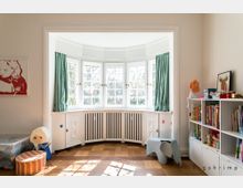 Helles Kinderzimmer mit einer großen Fensterfront im Erkerstil, weißen Fensterrahmen und grünen Vorhängen. Der Raum ist eingerichtet mit einem Bücherregal, einer Spielzeugtruhe, einem Kinderstuhl in Elefantenform und Holzboden.