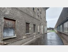 Eine schmale Gasse zwischen einem Gebäude mit grauer, rauer Fassade und teilweise zugemauerten oder mit Holzplatten verschlossenen Fenstern auf der linken Seite und einer langgestreckten Glasfront eines Industriegebäudes auf der rechten Seite. Der Boden ist regennass, und im Hintergrund sind Höfe, kleine Gebäude und eine Kirchturmspitze sichtbar.