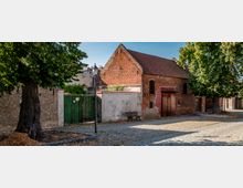 Kopfsteingepflasterte Straße in einem historischen Viertel mit einem niedrigen Backsteingebäude mit Satteldach, Holztür und kleinen Fenstern. Am Straßenrand stehen große Bäume, eine Laterne, eine Bank sowie eine grüne Holztorwand und angrenzende gemauerte Fassaden.