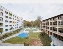Ein moderner Schulhof mit grünem Spielfeld, umliegenden Gehwegen und angelegten Grünflächen zwischen zwei Gebäuden: links ein Wohngebäude mit bunter Fassade und großen Fenstern, rechts ein Gebäude mit Backsteinoptik und Glasfenstern. Im Hintergrund Bäume und eine bewaldete Gegend.