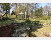 Ein Garten mit einer kleinen, runden Metallpavillon-Struktur im Hintergrund, umgeben von Bäumen und Sträuchern. Im Vordergrund befinden sich eine alte Steintreppe und verwilderte Grasflächen mit vereinzelt liegenden Steinen.
