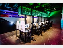 Ein moderner Gaming- oder E-Sport-Raum mit mehreren Schreibtischen, die jeweils mit Monitoren, Tastaturen, Mäusen und Gaming-Stühlen ausgestattet sind. Die Decke zeigt sichtbare Leitungen und Beleuchtungselemente in Grün- und Blautönen, die dem Raum ein technisches Ambiente verleihen.