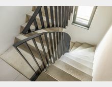 Eine Treppe aus hellem Beton mit schwarzem Metallgeländer, die spiralförmig nach unten verläuft. Im Hintergrund befindet sich ein Fenster, das Tageslicht in den Treppenbereich lässt.