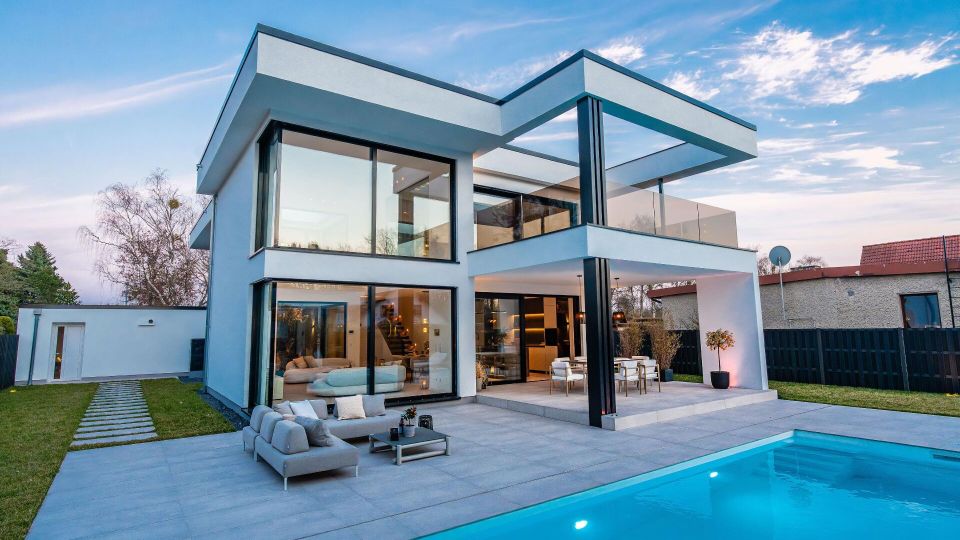 Modernes, zweistöckiges Wohnhaus mit Flachdach, großen bodentiefen Glasfronten und überdachter Terrasse mit schwarzen Stützen. Im Vordergrund liegt ein rechteckiger Swimmingpool mit heller Steinfläche, daneben Sitzmöbel; im Hintergrund sind Garten, Zaun und Nachbarhäuser zu sehen.