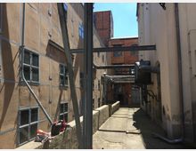 Ein enger Industriehof mit verwinkelten Gebäuden, teils aus Backstein und teils mit Putzfassaden. Sichtbare Rohre und Lüftungselemente verlaufen entlang der Wände, während kleine Fenster und ein schmaler Weg mit Schienen den Bereich prägen.