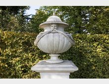 Eine steinerne Skulptur in Form einer großen, verzierten Vase mit einem menschlichen Gesicht und detaillierten Drapierungen, auf einem Podest stehend, umgeben von dichten grünen Hecken und Bäumen.