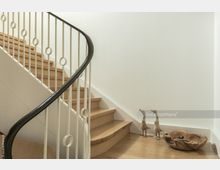 Holztreppe mit einem weißen, dekorativen Metallgeländer und geschwungenem schwarzem Handlauf, daneben eine helle Wand mit zwei hölzernen Gänsefiguren und einer rustikalen Holzschale auf dem Boden.