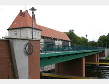 Brücke mit grünem Geländer und Backsteinpfeilern, daneben ein Gebäude mit hellem Putz und rotem Ziegeldach; im Hintergrund sind Bäume und ein bewölkter Himmel zu sehen.