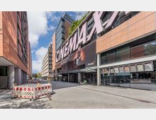 Straßenszene mit einem Gebäude, das ein großes Kino mit der Aufschrift "CinemaxX" zeigt. Das moderne Gebäude ist mit orangefarbenen Fassadenplatten verkleidet, im Vordergrund sind Absperrungen zu sehen, die eine Baustelle kennzeichnen.