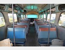 Innenraum eines alten Busses mit zwei Reihen von blauen und braunen Sitzen, die durch einen schmalen Mittelgang getrennt sind. Die großen Fenster an den Seiten lassen Tageslicht herein, während das abgenutzte Design auf das Alter des Fahrzeugs hinweist.