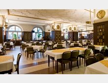 Innenraum eines Restaurants mit klassischen Holzmöbeln, gedeckten Tischen und gepolsterten Stühlen. Große Rundbogenfenster lassen Tageslicht herein, während dekorative Deckenverzierungen und dunkle Holzwände mit Regalen den Raum schmücken.