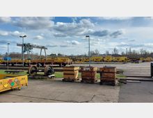 Ein Außenbereich eines Bahnbetriebswerks mit mehreren gelben Schienendienstfahrzeugen auf Gleisen, einem großen Metallkran und diversen Lagerkisten im Vordergrund. Im Hintergrund sind Bäume und vereinzelte Gebäude unter einem leicht bewölkten Himmel sichtbar.