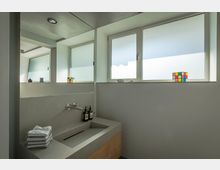 Modernes Badezimmer mit einem rechteckigen Waschbecken aus Stein, darüber ein großer Wandspiegel und eine minimalistische Armatur. Im Hintergrund ein Fenster mit mattem Glas, auf der Fensterbank zwei Rubik's Cubes und kleine Dekorationsgegenstände.