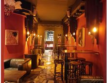 Ein schmaler Raum einer Bar mit warmem Licht, roten Wänden und abstrakten Bildern. Auf beiden Seiten befinden sich Sitzgruppen mit Ledersesseln, hohen Tischen und Hockern, im Hintergrund ist eine Theke mit Spiegeln und Flaschen sichtbar.