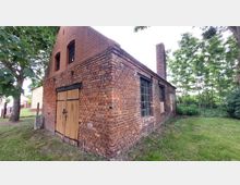 Backsteingebäude mit einfachem Satteldach und Schornstein, größtenteils aus roten Ziegeln errichtet, mit großen, rechteckigen Fenstern und einer Holztür auf der Vorderseite; umgeben von einer Wiese und Bäumen.