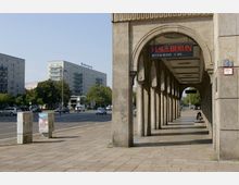 Arkaden eines Gebäudes mit Säulen auf der rechten Seite, darüber ein Schild mit der Aufschrift „Haus Berlin Restaurant • Café“. Im Hintergrund Gebäude im typischen Baustil der DDR-Architektur, mit einer breiten Straße und parkenden sowie fahrenden Autos.