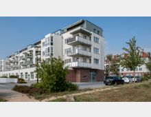 Mehrfamilienhaus in moderner Bauweise mit vier Stockwerken und Balkonen, kombiniert aus weiß verputzter und roter Backsteinfassade. Im Vordergrund eine begrünte Fläche, am Rand parken mehrere Autos, und der Himmel ist klar.