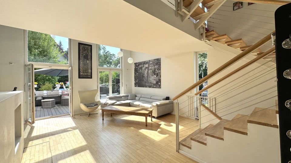 Heller, moderner Wohnbereich mit doppelter Raumhöhe, großen Fensterfronten und offener Treppe mit Holzstufen und Seilgeländer zur Galerie. Links führt eine Glastür auf eine Terrasse, im Raum stehen ein Ecksofa, ein Sessel und ein Couchtisch auf hellem Holzboden.