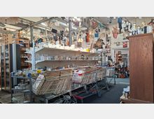 Innenansicht eines vollgestellten Trödelladens in einer hohen, industriellen Halle mit Metallstützen und vielen von der Decke hängenden Lampen und Leuchten. Regale und Tische sind dicht mit Gläsern, Geschirr und Kleinteilen gefüllt; davor stehen Einkaufswagen mit gestapelten Schallplatten.