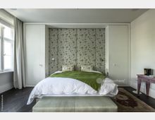 Schlafzimmer mit Doppelbett vor einer dekorativen Rückwand mit floralen Mustern. Links und rechts des Bettes befinden sich weiße Einbauschränke, daneben ist ein großes Fenster mit Vorhängen sichtbar; der Boden ist aus dunklem Holz.