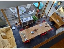 Innenraum einer modernen Wohn- und Essbereich-Kombination mit großen Glasfenstern, die auf eine Holzterrasse führen. Der Raum enthält einen Holztisch mit gemischten Stühlen, ein gelbes Sofa, ein weißes Sideboard und Dekorationen wie Pflanzen, Bilder und Kerzen.