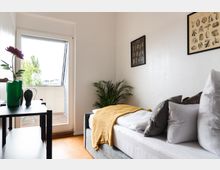 Helles Schlafzimmer mit Einzelbett, dekoriert mit farbigen Kissen und einer gelben Decke; im Hintergrund ein Balkon mit Glastür, Pflanzen sowie zwei Poster an der weißen Wand. Ein kleines Regal mit Dekorationsobjekten steht links im Raum.