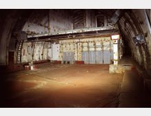 Ehemaliger unterirdischer Bunkerraum mit gewölbter Decke, verrosteten Metallteilen und abgenutzten Wänden; der Boden zeigt Anzeichen von Verfall, und Kabel liegen verstreut. Die Umgebung wirkt verlassen und ungenutzt.