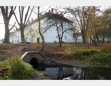 Mehrfamilienhaus aus der Nachkriegszeit, umgeben von Bäumen in einer herbstlichen Parklandschaft; im Vordergrund ein kleiner gemauerter Durchlass mit Wasserlauf.
