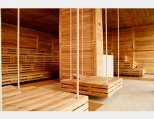 Holzverkleideter Wellnessbereich mit hölzernen Liegen und schwingenden Sitzflächen, die an Seilen von der Decke hängen. Der Raum hat eine warme, natürliche Atmosphäre mit einem Boden aus glattem Beton und durchgehender Holzverkleidung an Wänden und Decke.
