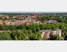 Luftaufnahme einer Stadt mit roten Ziegeldächern und historischen Gebäuden, umgeben von dichten grünen Bäumen. Im Hintergrund sind flache Landschaften und eine Kirche mit einem hohen Turm sichtbar.