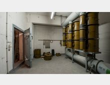 Ein Betonraum mit einer schweren, geöffneten Stahltür und gelb lackierten, zylindrischen Behältern, die auf Metallregalen montiert sind. An der Wand hängen Werkzeuge und Gurte, der Boden ist mit einzelnen Behältern und Rohren belegt, die Teil eines Belüftungssystems zu sein scheinen.