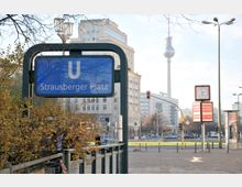 U-Bahn-Eingang mit blauem Schild „Strausberger Platz“ im Vordergrund, im Hintergrund der Berliner Fernsehturm, umliegende Gebäude, Ampeln und eine Uhr auf einem Pfosten. Laub bedeckt teilweise den unteren Bildrand.