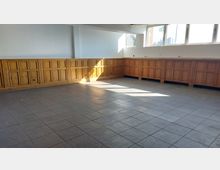 Leerer Raum mit Boden aus grauen Fliesen und Wänden, die teilweise mit holzvertäfelten Schränken ausgestattet sind. Das Tageslicht fällt durch mehrere große Fenster an der oberen Wand.