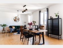 Ein modern eingerichtetes Wohnzimmer mit Essbereich, einem Holztisch und gemischten Stühlen. Im Hintergrund befinden sich ein grünes Sofa, ein runder Wandregalrahmen, ein schwarzer Schrank mit Glasschiebetüren sowie Pflanzen und Dekorationen, die den Raum akzentuieren; Tageslicht fällt durch große Fenster mit grauen Vorhängen.