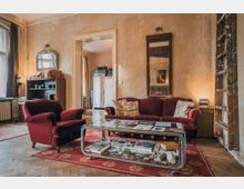 Wohnzimmer in einem Altbau mit abgenutzten Wänden, roten Samtmöbeln und einem gläsernen Couchtisch auf einem roten Teppich. Im Hintergrund sind eine geöffnete Tür, ein großer Spiegel, ein Regal und ein Schrank sichtbar.