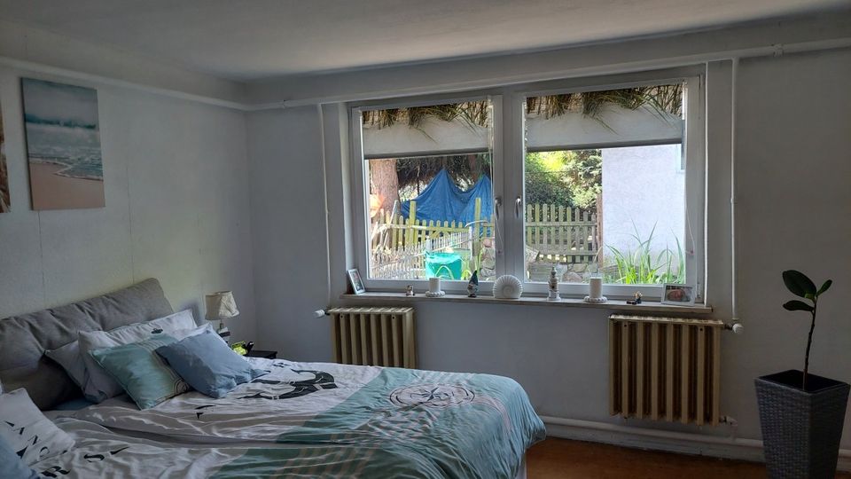 Schlafzimmer mit großem Doppelbett, hellen Wänden und Holzfußboden. Eine breite Fensterfront mit Fensterbankdekoration und zwei Heizkörpern blickt in einen kleinen Garten mit Zaun und Pflanzen.