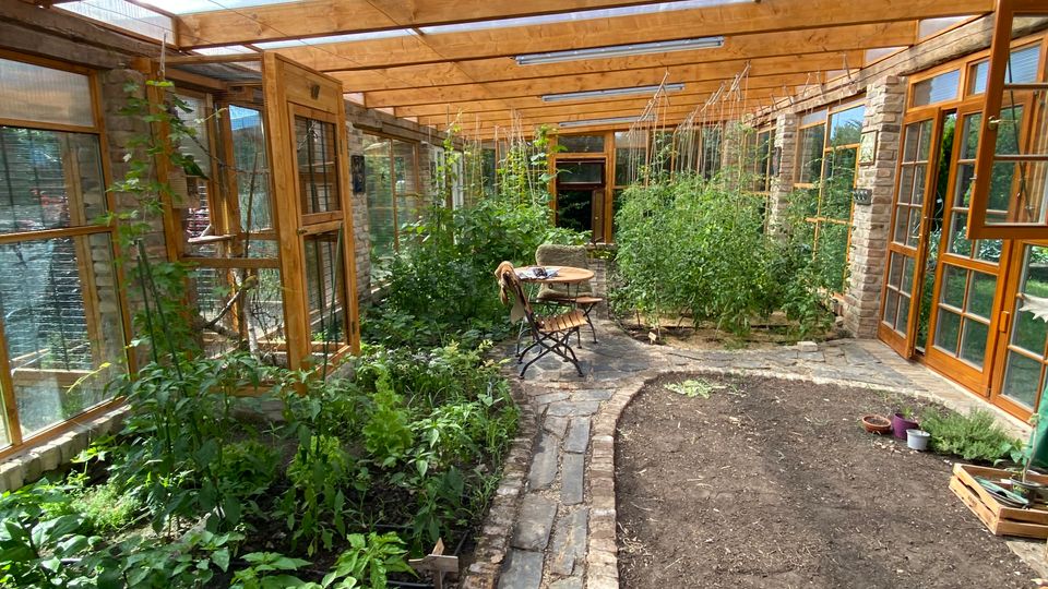 Überdachter Gewächshausgang mit Holzträgern und transparentem Dach, flankiert von Glasfenstern und Holztüren an einer Steinwand. Entlang eines gepflasterten Weges stehen dicht bepflanzte Beete mit Rankhilfen, daneben eine freie Erdfläche und ein kleiner Tisch mit Stühlen.