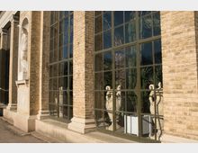 Fassade eines Gebäudes mit hellen Backsteinmauern, großen rechteckigen Fenstern und sichtbaren Pflanzen sowie dekorativen Statuen im Inneren. Links ist eine in die Wand eingelassene Steinstatue in einer Arkade zu sehen.