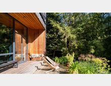 Holzterrasse eines modernen Hauses mit vertäfelter Holzfassade und großen Glasfenstern, umgeben von dichtem Wald. Auf der Terrasse stehen Liegestühle, Pflanzen in Töpfen und eine mit Fell bedeckte Sitzbank.