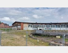 Altes Bahnbetriebswerk mit einem halbkreisförmigen Lokschuppen aus roten Ziegeln und großen Fensterfronten, teilweise mit Graffiti versehen. Im Vordergrund befindet sich eine stillgelegte Drehscheibe, umgeben von einem Bauzaun.