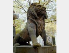 Steinstatue eines sitzenden Löwen auf einem Sockel, umgeben von Bäumen mit frischem Laub. Im Hintergrund sind unscharf ein Gebäude und Fenster zu sehen.