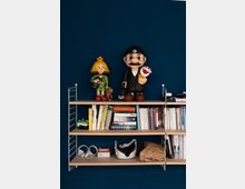 Ein Wandregal vor einer dunkelblauen Wand, mit mehreren Ebenen gefüllt: Oben befinden sich große Figuren, darunter Bücher, ein grüner Controller, Kopfhörer und eine Virtual-Reality-Brille.