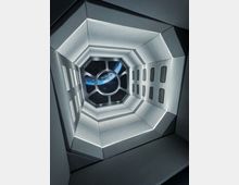 Futuristischer, enger Korridor mit weißen, beleuchteten Wänden und rechteckigen Lichtfeldern; am Ende ein Fenster mit Blick auf die Erde im Weltraum.